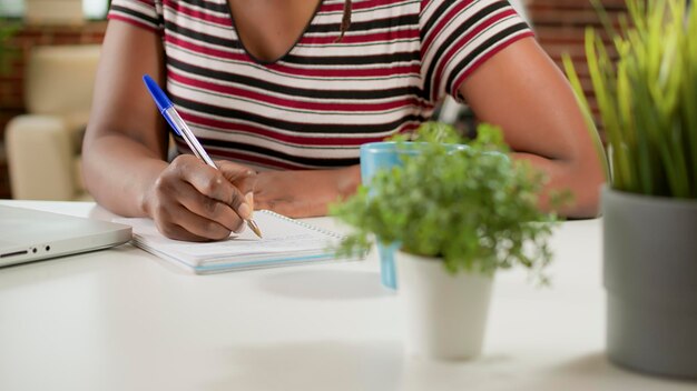 Mujer independiente tomando notas en el seminario web de la escuela en línea, aprendiendo sobre negocios y haciendo trabajo remoto. Escribiendo en un cuaderno y usando un portátil para asistir a clase, trabajando de forma remota desde casa. De cerca.