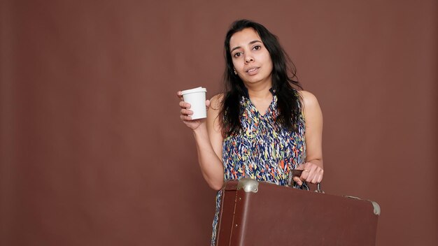 Mujer india preparándose para viajar de vacaciones con una maleta y una taza de café en la mano. Sentirse feliz de irse de vacaciones con equipaje y bebida.