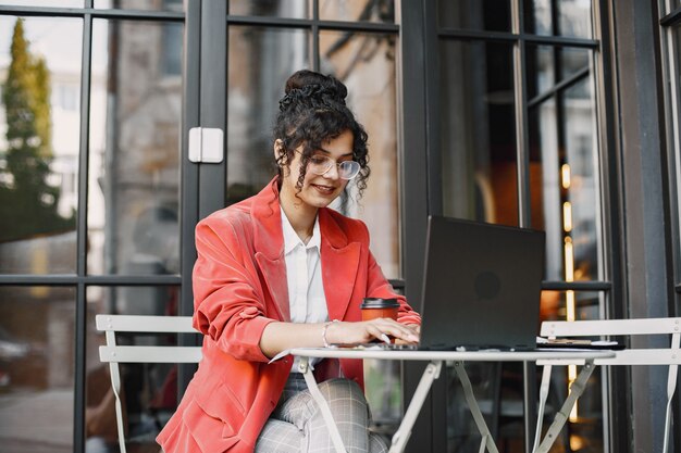 Mujer india trabajando en un portátil en un café de la calle. Usar ropa elegante e inteligente: chaqueta, gafas