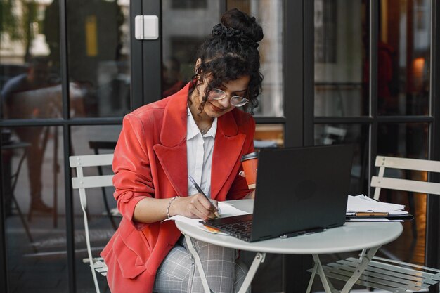 Mujer india trabajando en un portátil en un café de la calle. Usar ropa elegante e inteligente: chaqueta, gafas