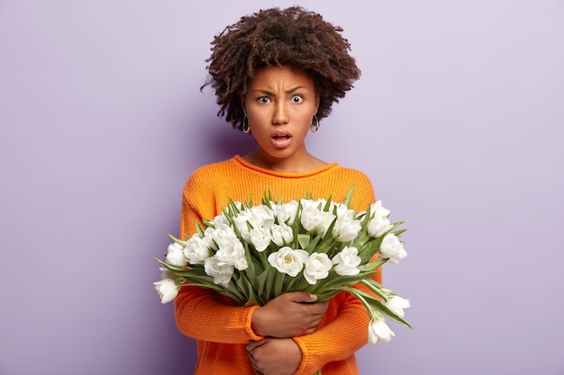 La mujer indignada e insatisfecha se ve enojada, sostiene flores blancas, usa un jersey casual naranja, modela sobre una pared púrpura, expresa emociones negativas, escucha malas noticias. Mujer con tulipanes
