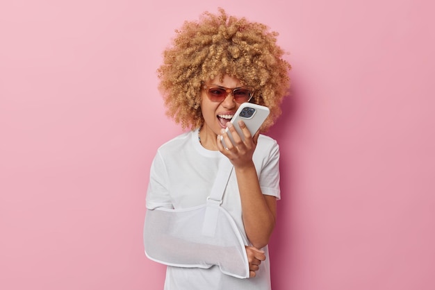 Una mujer indignada grita enojada y se queja de que el teléfono móvil lee algo molesto que fue arrojado por un mensaje tiene un brazo roto, usa gafas de sol y una camiseta blanca se encuentra en el interior contra un fondo rosa