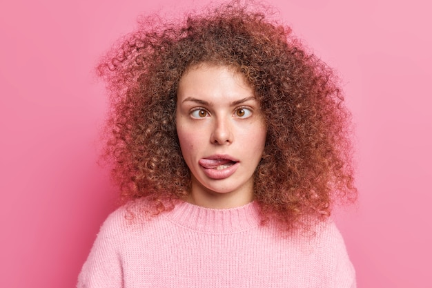 La mujer infantil de pelo rizado divertido hace muecas cruza los ojos y saca la lengua muestra caras divertidas que intenta entretener a un amigo vestido con un jersey casual aislado sobre una pared rosa.