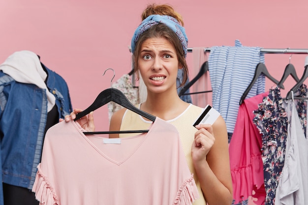 Foto gratuita mujer infeliz haciendo compras, parada en una tienda de ropa, sosteniendo un vestido nuevo y una tarjeta de crédito, recibiendo una inyección de dinero, teniendo una crisis financiera, queriendo comprar ropa nueva de inmediato. extensiones de compras