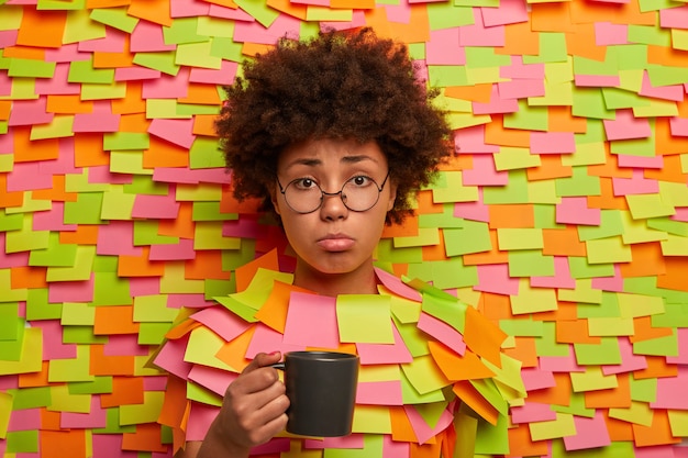 Mujer infeliz insatisfecha sostiene una taza de té, se siente cansada del trabajo, descontenta por tener algunos problemas, tiene expresión de lástima, asoma la cabeza en la pared de papel con pegatinas de colores. Estudiante de fatiga