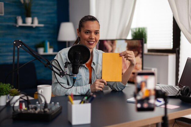 Mujer influyente que presenta un libro amarillo frente a un teléfono inteligente de grabación. Simulación de la revisión de transmisión del creador de contenido para el canal de medios sociales en un estudio doméstico con micrófono y computadora portátil.