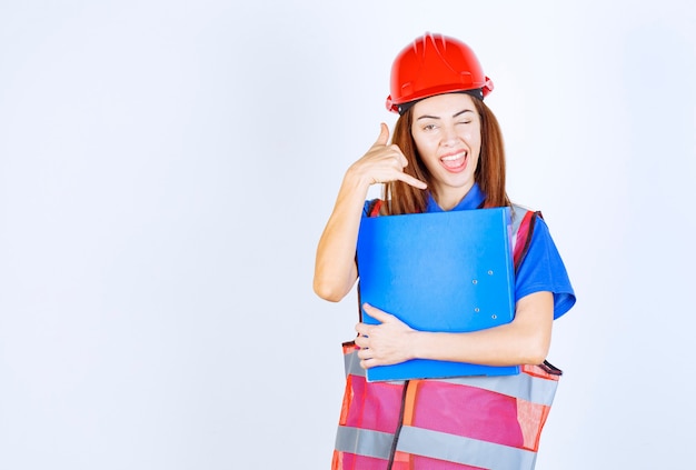 Mujer ingeniera en casco rojo sosteniendo una carpeta de proyecto azul y pidiendo una llamada.