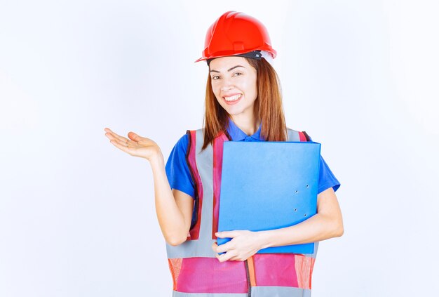 Mujer ingeniera en casco rojo sosteniendo una carpeta de proyecto azul y presentando a alguien o algo.