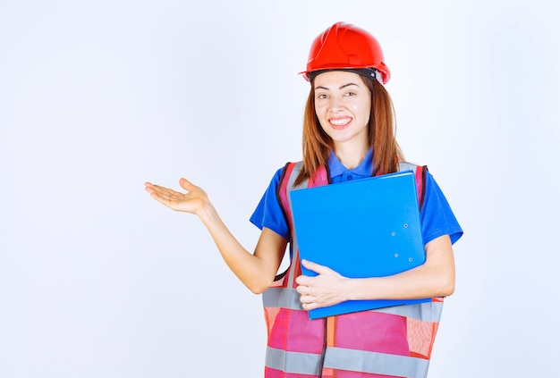 Mujer ingeniera en casco rojo sosteniendo una carpeta de proyecto azul y presentando a alguien o algo.