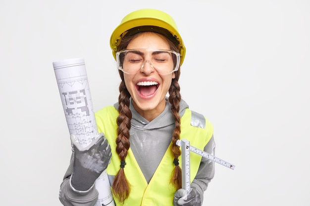 Mujer ingeniera llena de alegría se ríe felizmente mantiene los ojos cerrados se divierte sostiene el proyecto arquitectónico y la cinta métrica se regocija para lograr excelentes resultados vestida con uniforme de trabajo aislado en una pared blanca