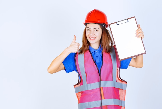 Foto gratuita mujer ingeniera en uniforme sosteniendo un archivo de informes en blanco y mostrando el signo de satisfacción.