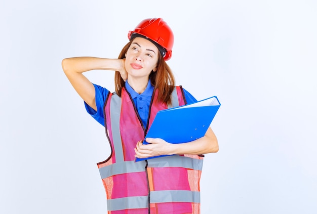 Mujer ingeniero en casco rojo sosteniendo una carpeta de proyecto azul y pensando.