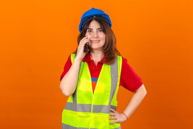 Mujer ingeniero en construcción chaleco y casco de seguridad hablando por teléfono móvil sonriendo alegremente de pie sobre la pared naranja aislada