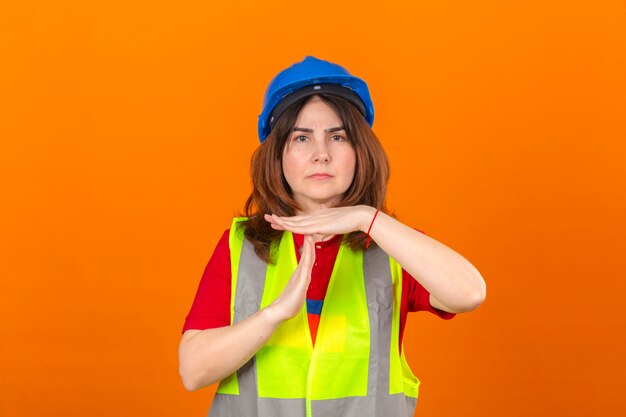 Mujer ingeniero vistiendo chaleco de construcción y casco de seguridad haciendo gesto de tiempo fuera con las manos frustradas y cara seria sobre la pared naranja aislada