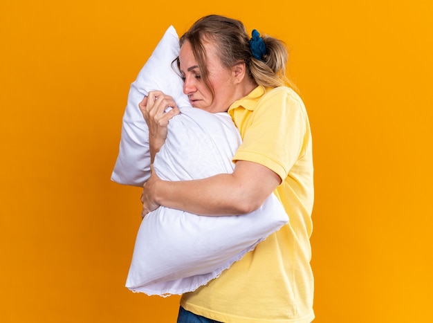 Mujer insalubre en camisa amarilla malestar que sufre de gripe y frío abrazando la almohada de pie sobre la pared naranja