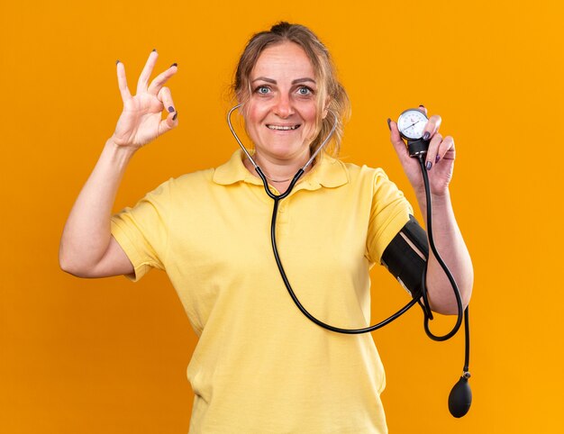 Mujer insalubre en camisa amarilla que se siente mejor midiendo su presión arterial con tonómetro sonriendo mostrando el signo de ok de pie sobre la pared naranja