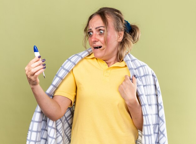 Foto gratuita mujer insalubre envuelta en una manta sintiéndose terrible sufrimiento de gripe y termómetro de retención fría mirando asustada