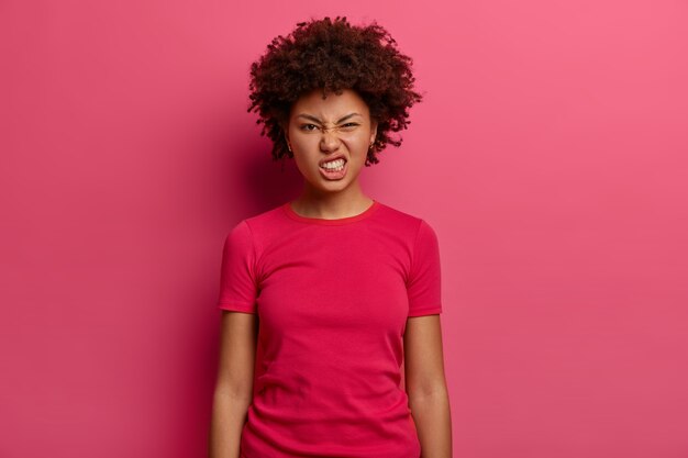 Mujer insatisfecha molesta aprieta los dientes y se siente irritada, parece disgustada, vestida con una camiseta informal, posa en el interior contra la pared rosa. Concepto de expresiones faciales negativas