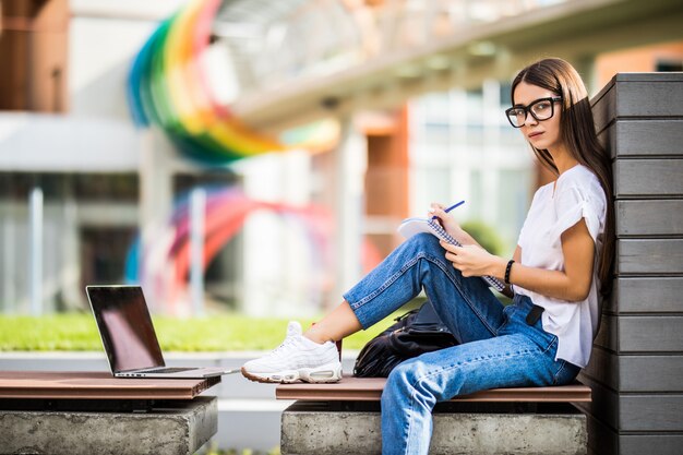 Mujer inteligente con gafas haciendo notas en el planificador y navegando portátil mientras está sentado en el banco en el patio del edificio moderno en la ciudad
