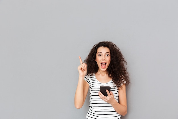 Mujer inteligente con teléfono móvil en la mano manteniendo el dedo índice apuntando hacia arriba expresando que tiene una idea o diciendo eureka sobre una pared gris