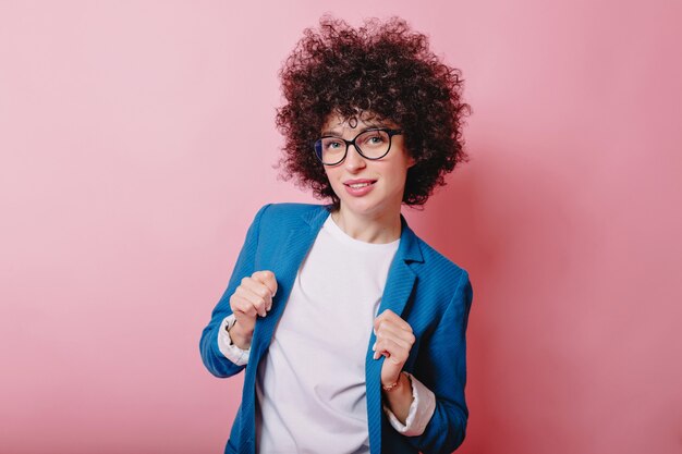 Mujer inteligente vestida con chaqueta azul y gafas posa en rosa con una sonrisa encantadora.