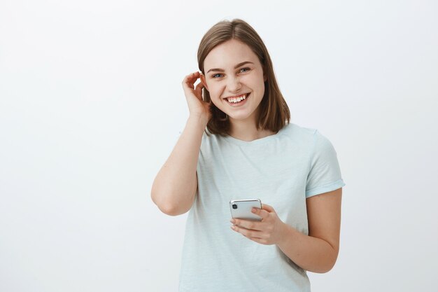 Mujer intercambiando números con chico lindo en festival. Encantadora chica joven de aspecto amistoso moviendo el cabello detrás de la oreja coqueta y riendo mientras mira sosteniendo el teléfono inteligente contra la pared gris