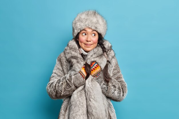 Mujer inuit pensativa vestida con sombrero de piel gris y guantes de punto de abrigo viste ropa de invierno cálida aislada sobre pared azul de estudio