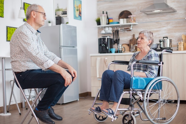 Mujer inválida jubilada en silla de ruedas que tiene una conversación con el anciano marido en la cocina. Anciano hablando con esposa. Vivir con una persona discapacitada con discapacidad para caminar
