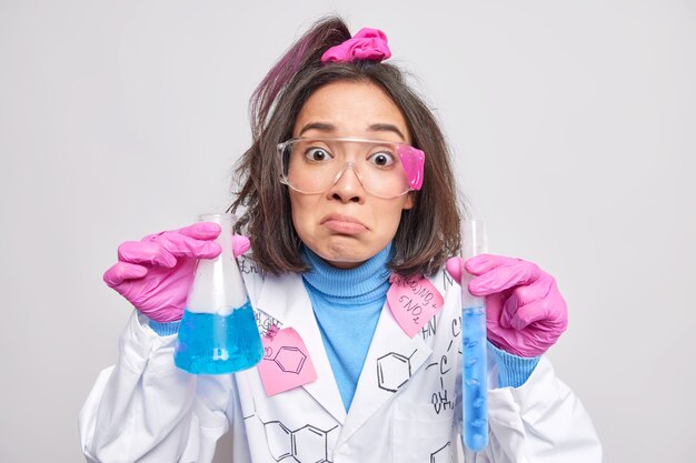 Mujer investigadora química sostiene líquido azul en matraces se siente disgustado usa gafas protectoras bata blanca guantes de goma aislados en gris