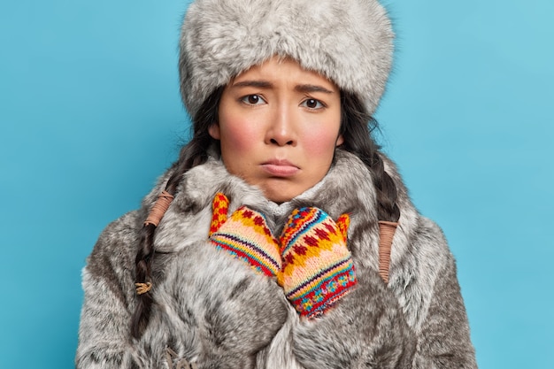 Mujer de invierno mira tristemente al frente siente frío vestida con gorro de piel gris y abrigo con guantes de punto cálidos vestidos para el clima invernal aislado sobre una pared azul