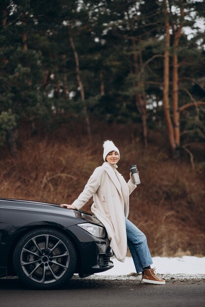 Mujer en invierno sentada en el capó del auto y bebiendo café