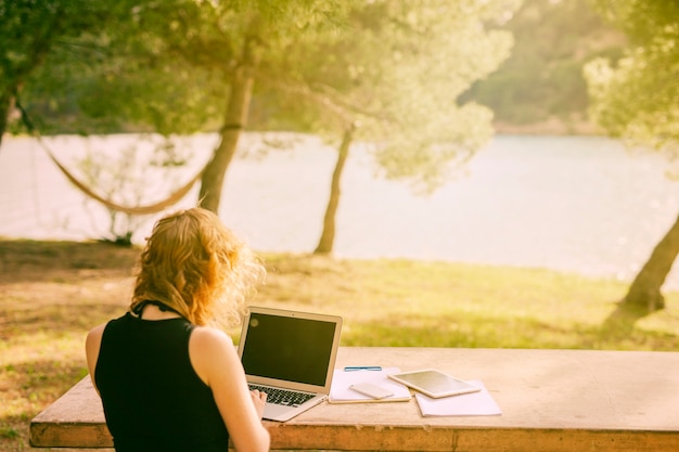 Mujer irreconocible que se sienta en el escritorio y que trabaja en la computadora portátil en naturaleza