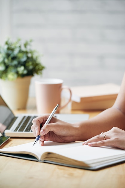 Mujer irreconocible sentada en el escritorio en el interior y escribiendo en planificador