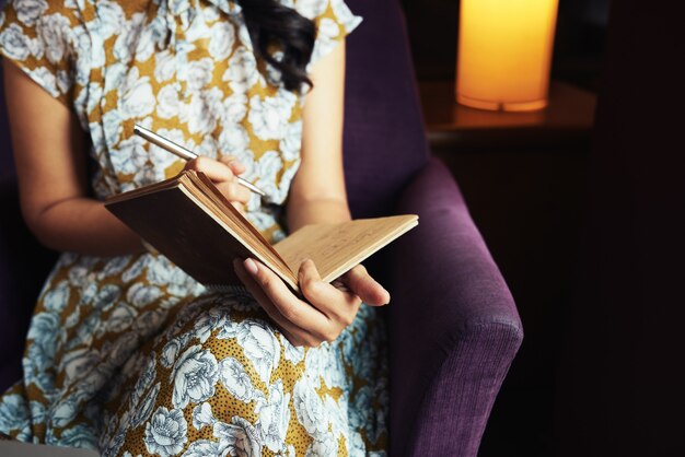 Mujer irreconocible sentada en un sillón y escribiendo en el diario