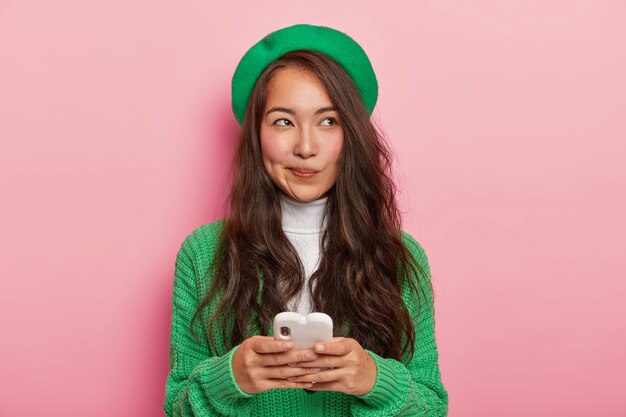 La mujer japonesa de pelo bastante oscuro utiliza un teléfono celular moderno para enviar mensajes de texto, navega por Internet, tiene una expresión pensativa