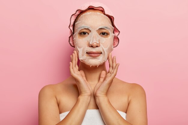 Mujer japonesa seria pone mascarilla nutritiva en la cara, aplica producto humectante en la piel, usa gorro de baño, posa contra la pared rosada. Concepto de tratamiento de feminidad, cosmetología y spa.