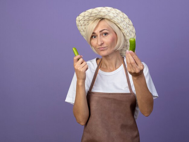 Mujer jardinero rubia de mediana edad complacida en uniforme con sombrero sosteniendo mitades de pimiento aislado en la pared púrpura con espacio de copia