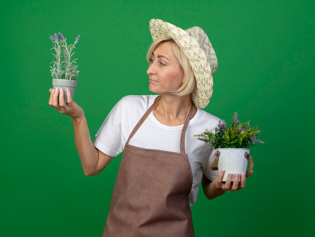 Mujer jardinero rubia de mediana edad confundida en uniforme con sombrero sosteniendo macetas mirando uno de ellos aislado en la pared verde