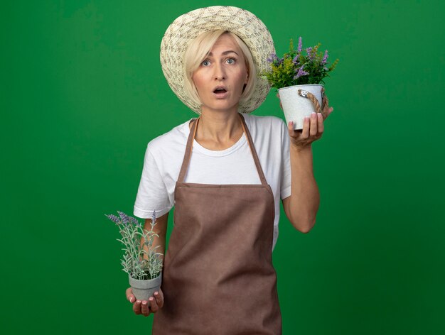 Mujer jardinero rubia de mediana edad sorprendida en uniforme con sombrero sosteniendo macetas mirando hacia arriba