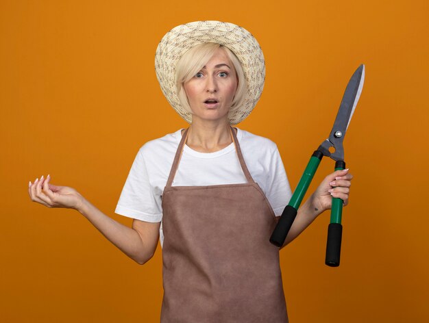 Mujer jardinero rubia de mediana edad sorprendida en uniforme con sombrero sosteniendo tijeras de podar mostrando la mano vacía aislada en la pared naranja
