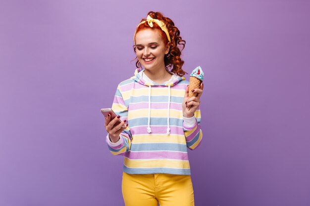 Mujer de jengibre en aderezo de cabello amarillo con sonrisa está charlando en el teléfono inteligente y sosteniendo helado azul