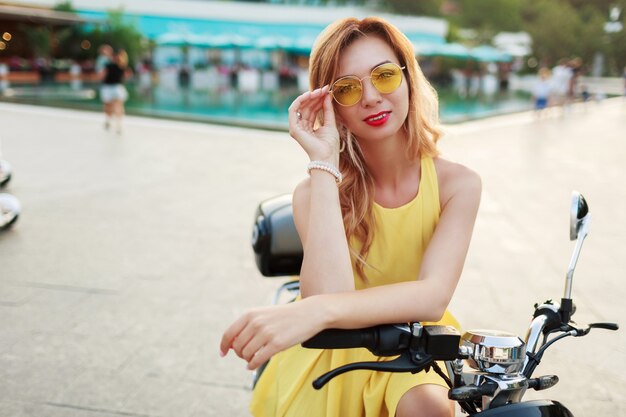 Mujer de jengibre alegre en gafas amarillas y vestido vintage sentado en una elegante moto. Estado de ánimo de vacaciones. Día soleado de verano. Labios rojos.