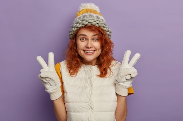 La mujer de jengibre complacida sonríe agradablemente, levanta las manos y hace un gesto de paz, usa gorro de invierno, guantes y chaleco blanco, está de buen humor, aislado sobre fondo púrpura.