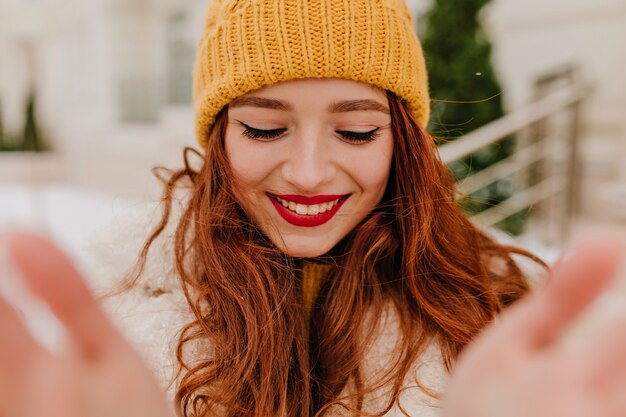 Mujer de jengibre emocionada haciendo selfie al aire libre con una sonrisa sincera. Chica fascinante con pelo largo rojo disfrutando del invierno.