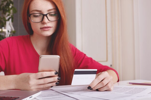 Mujer de jengibre con gafas, revisa su cuenta en un teléfono inteligente, tiene tarjeta de crédito, tiene un descanso después del trabajo con una computadora portátil