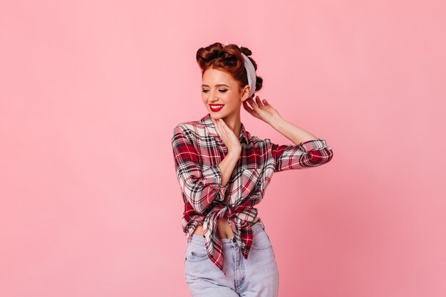 Mujer de jengibre glamorosa posando en el espacio rosa. Disparo de estudio de chica pinup soñadora en camisa a cuadros.