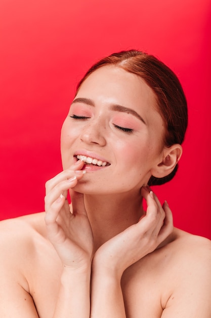 Mujer de jengibre inspirada tocando los labios. Foto de estudio de modelo femenino desnudo posando con los ojos cerrados sobre fondo rojo.