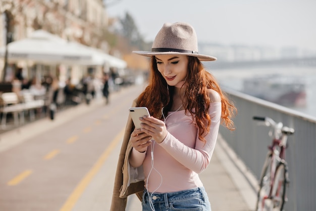 Mujer de jengibre interesada con teléfono en manos comprobando el correo en la muralla de la ciudad