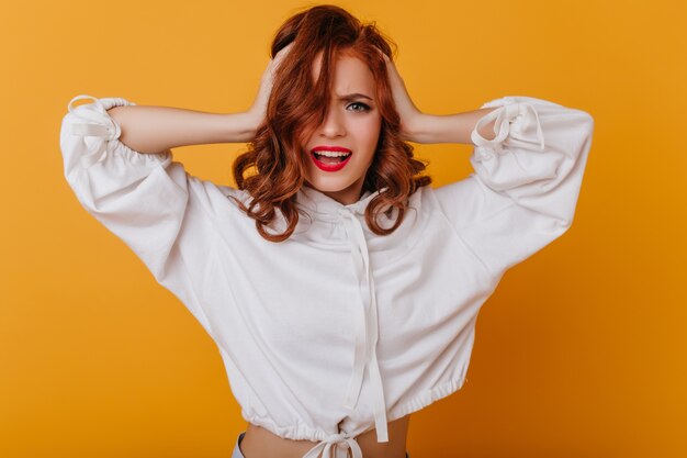 Mujer de jengibre preocupada con labios rojos posando en la pared amarilla. Modelo de mujer emocional en suéter blanco tocando su cabello.