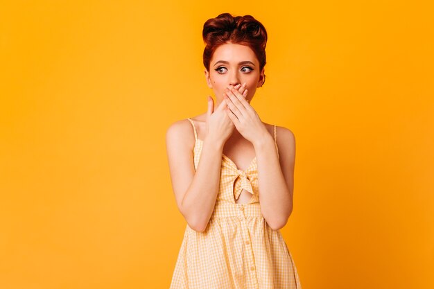 Mujer de jengibre sensual que cubre la boca con las manos. Foto de estudio de moda modelo femenino pinup aislado en espacio amarillo.